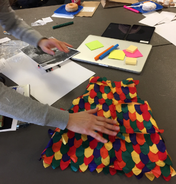 Multicoloured textured child’s dress lying on a table amongst digital tablets, pads of paper, sticky notes and markers. The dress is being touched by a human hand.