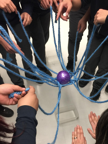 A web is formed by several long ropes intersecting in the middle. Students hold the ends of the ropes and together move a ball siting in the middle of the web into a container.