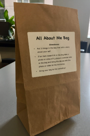 Paper bag sitting on a table with a sticker on the front that reads: All About Me Bag, Directions: Put 3 things in the bag that tells a story about yourself. If an item doesn’t fit in the bag take a photo or video of it, place a reminder note in the bag and bring the device with the photo or video to the workshop. Bring your bag to the workshop!